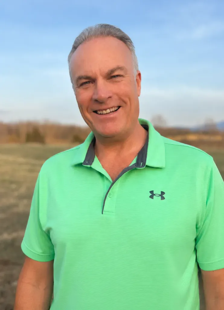 Smiling man wearing a bright green polo shirt standing outdoors in an open field at sunset.