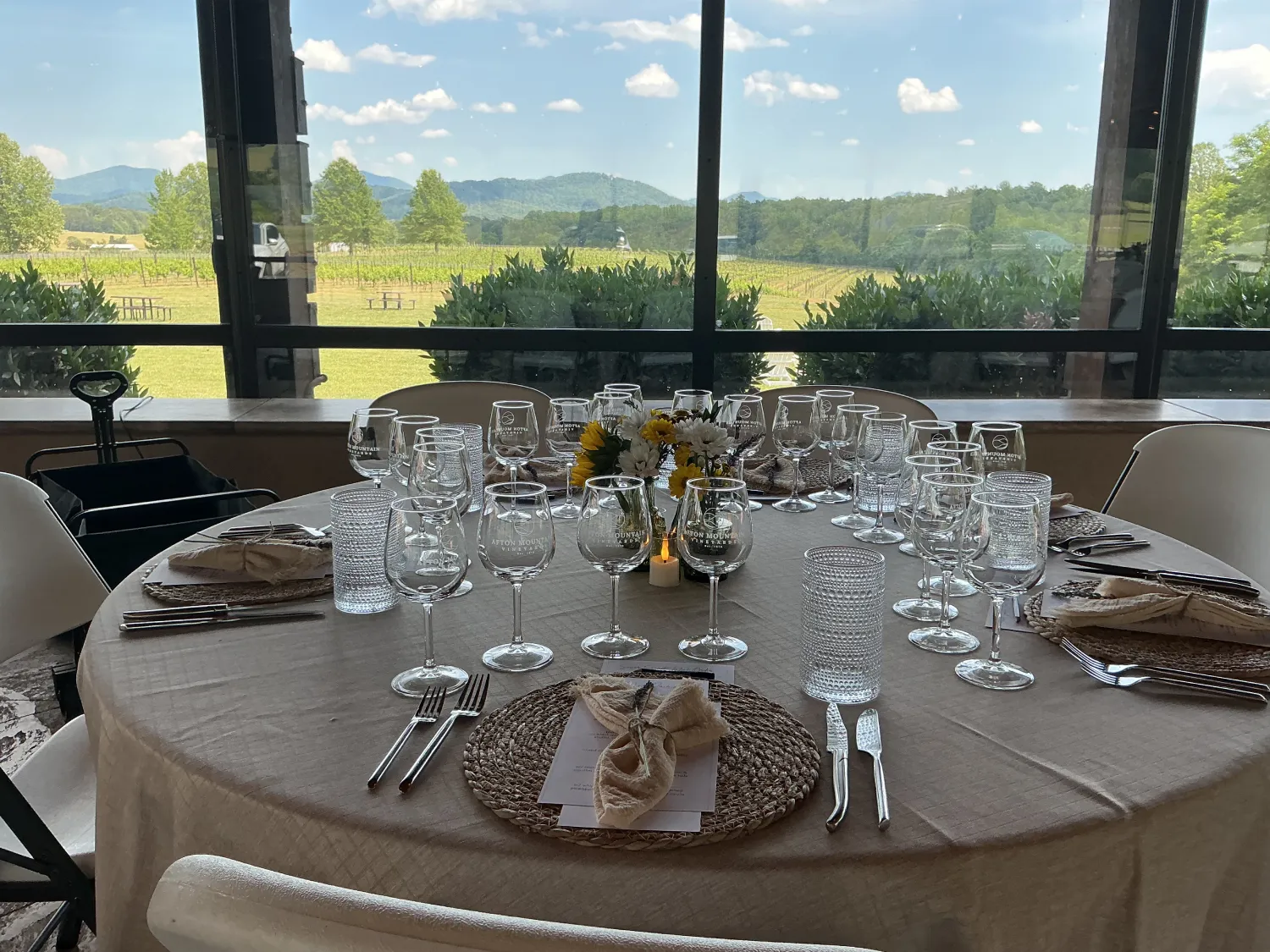 Elegantly set round dining table with wine glasses, woven placemats, and floral accents positioned beside large windows overlooking vineyard rows and distant mountains.