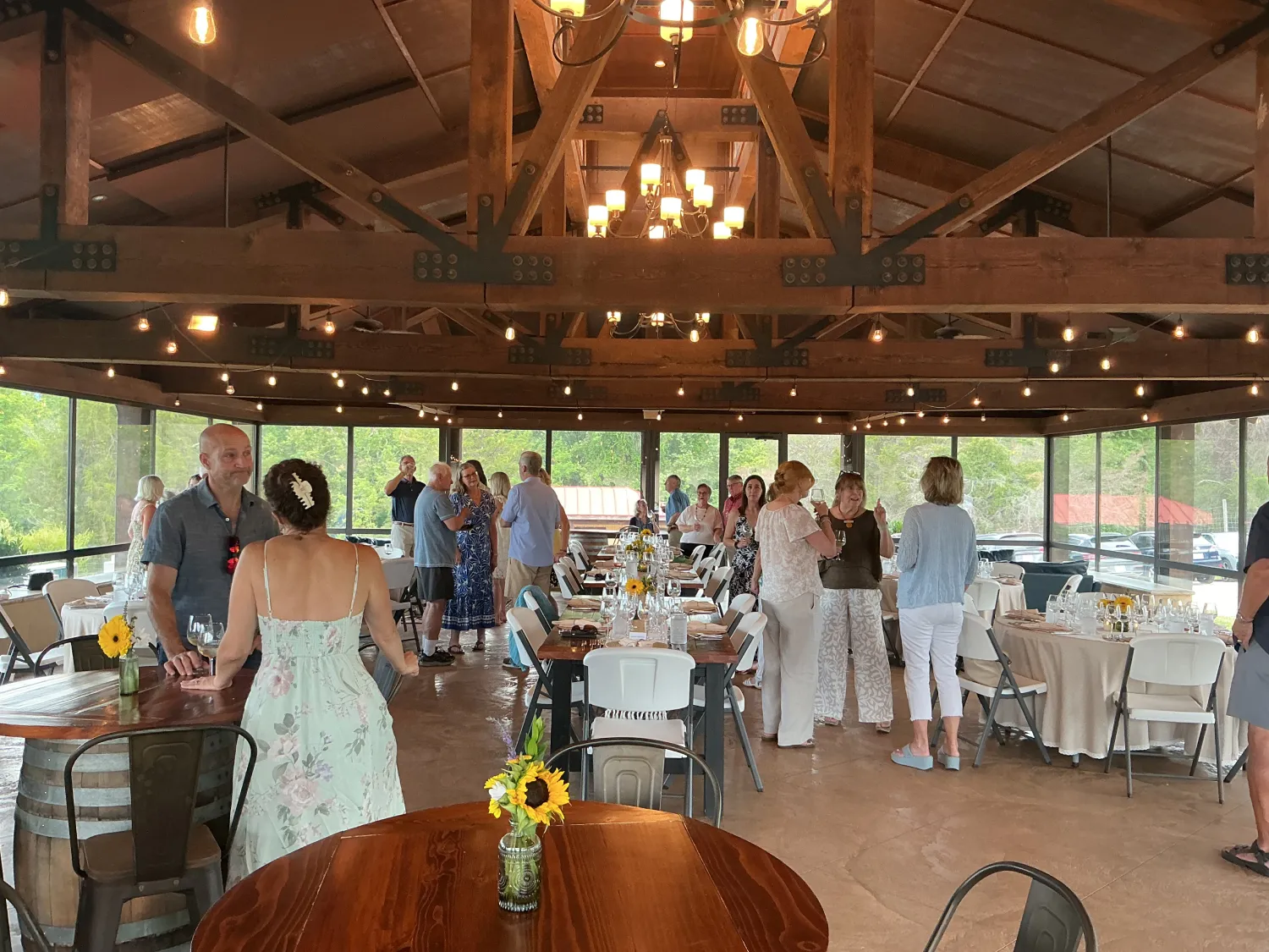 Guests mingle and socialize inside a rustic vineyard event pavilion featuring exposed timber beams, string lights, and panoramic views of the surrounding countryside.