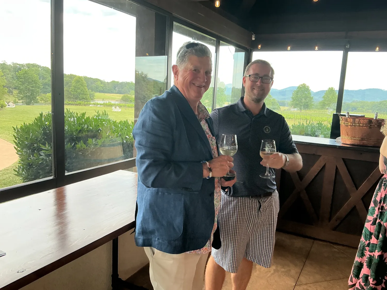 Two guests holding wine glasses and smiling during a vineyard gathering, standing beside large windows overlooking rolling vineyards and mountain scenery.