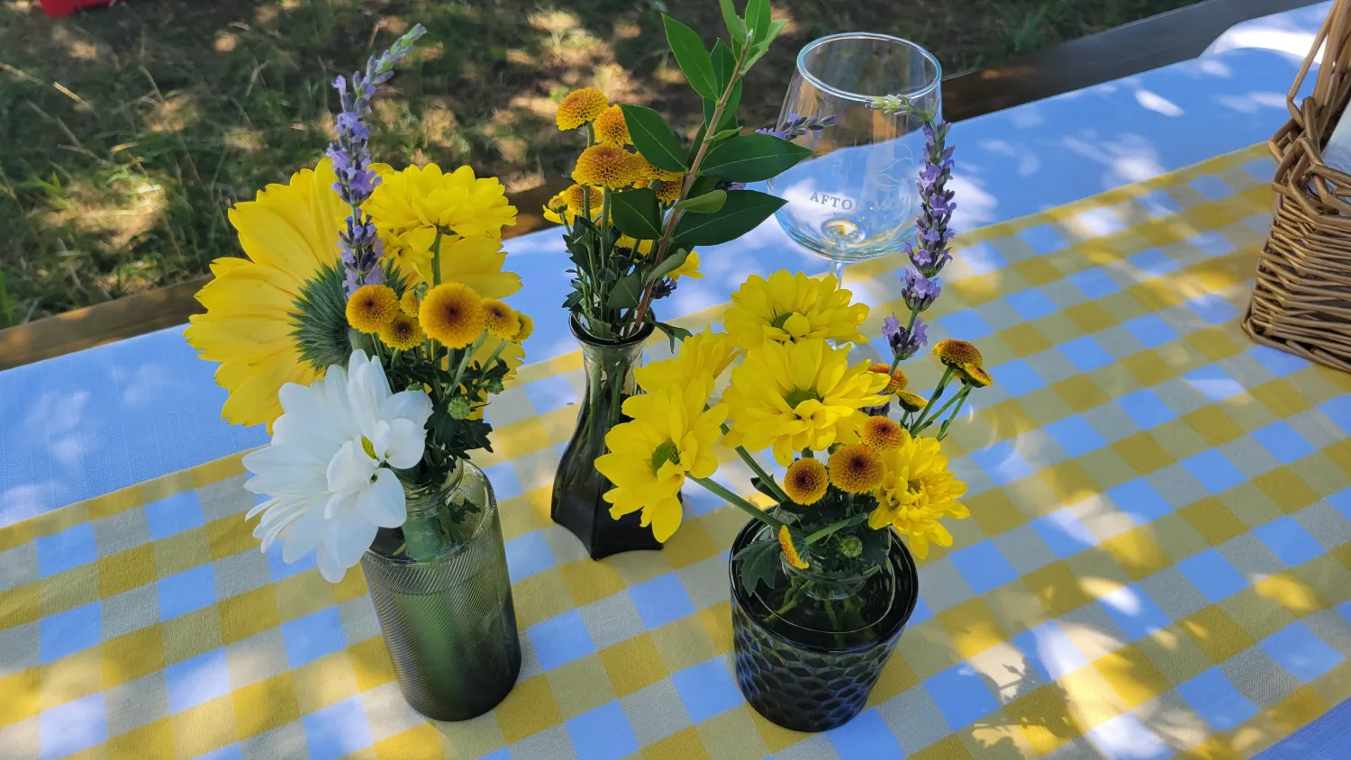 Fresh floral centerpieces featuring yellow, white, and lavender blooms arranged in glass vases atop a yellow-and-white gingham tablecloth for an outdoor vineyard picnic.